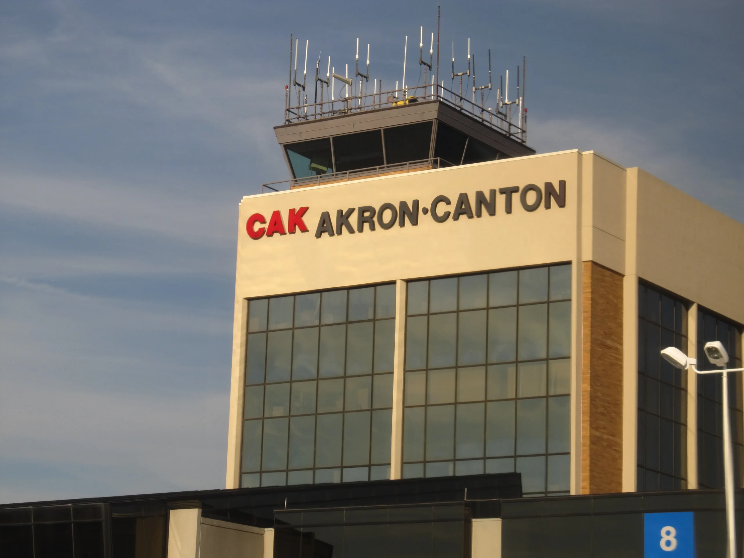Low-angle view of the Akron-Canton Airport (CAK) main terminal building, showing large dark-tinted glass windows, building signage, and the air traffic control tower equipped with communication antennas against a blue sky.