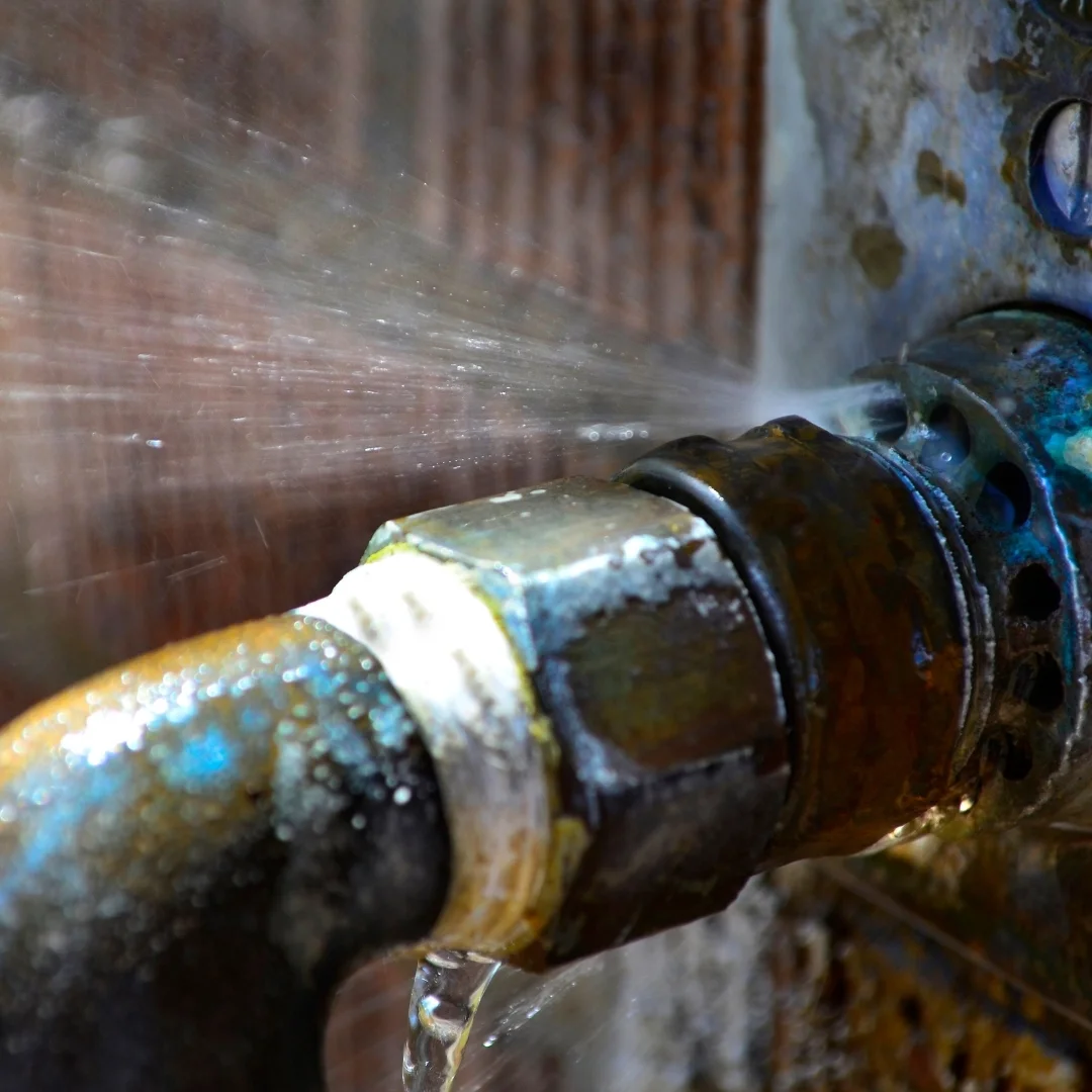Extreme close-up of water spraying forcefully from a burst pipe fitting and leaking from a corroded metal connection, illustrating an emergency plumbing failure.