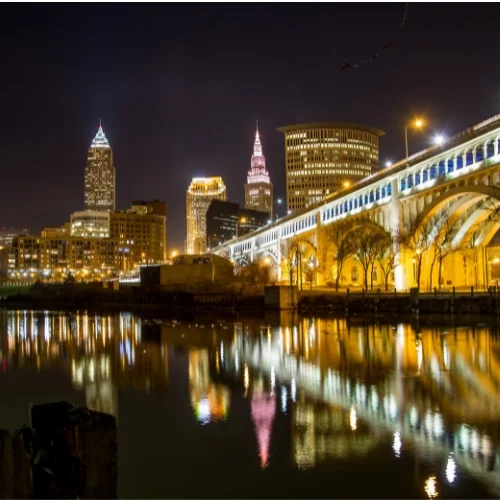 Nighttime view of the Cleveland, Ohio city skyline reflecting across the Cuyahoga River, featuring the illuminated Key Tower, Terminal Tower, and the lit arches of the Detroit-Superior Bridge.