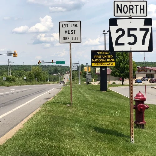 A roadside view along a highway showing a Pennsylvania State Route 257 North sign and a red fire hydrant in the foreground. In the background, signs for "Left Lane Must Turn Left" and a "First United National Bank Drive-In ATM" stand along a grassy median under a partly cloudy sky.