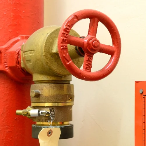 Close-up of a brass fire hose angle valve connected to a red vertical standpipe riser, featuring a red handwheel marked "OPEN" and a security seal with a paper inspection tag.