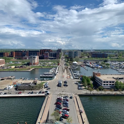 High-angle aerial view looking south down State Street from the Bicentennial Tower in Erie, Pennsylvania. The scene overlooks the Donjon Shipbuilding slips, the east and west canal basins filled with docked boats, and downtown city buildings under a bright sky with scattered clouds.