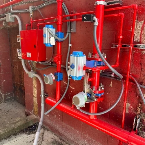 Outdoor fire protection piping system featuring red metal headers, electronic control boxes, grey electrical conduit, and blue motorized control valves mounted against a red brick wall.
