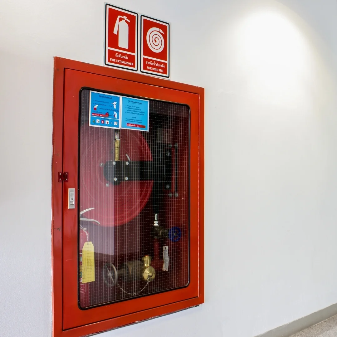A wall-mounted red fire protection cabinet with a wire-glass door containing a fire hose reel, valve, and a fire extinguisher with a yellow inspection tag.