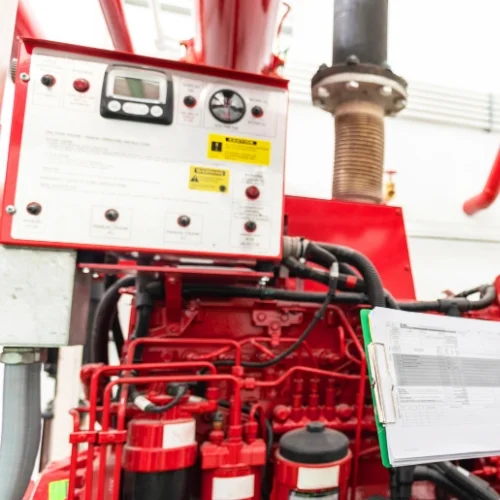 Close-up of a red diesel fire pump driver engine control panel and machinery, featuring mechanical piping, gauges, and an inspection checklist on a green clipboard.