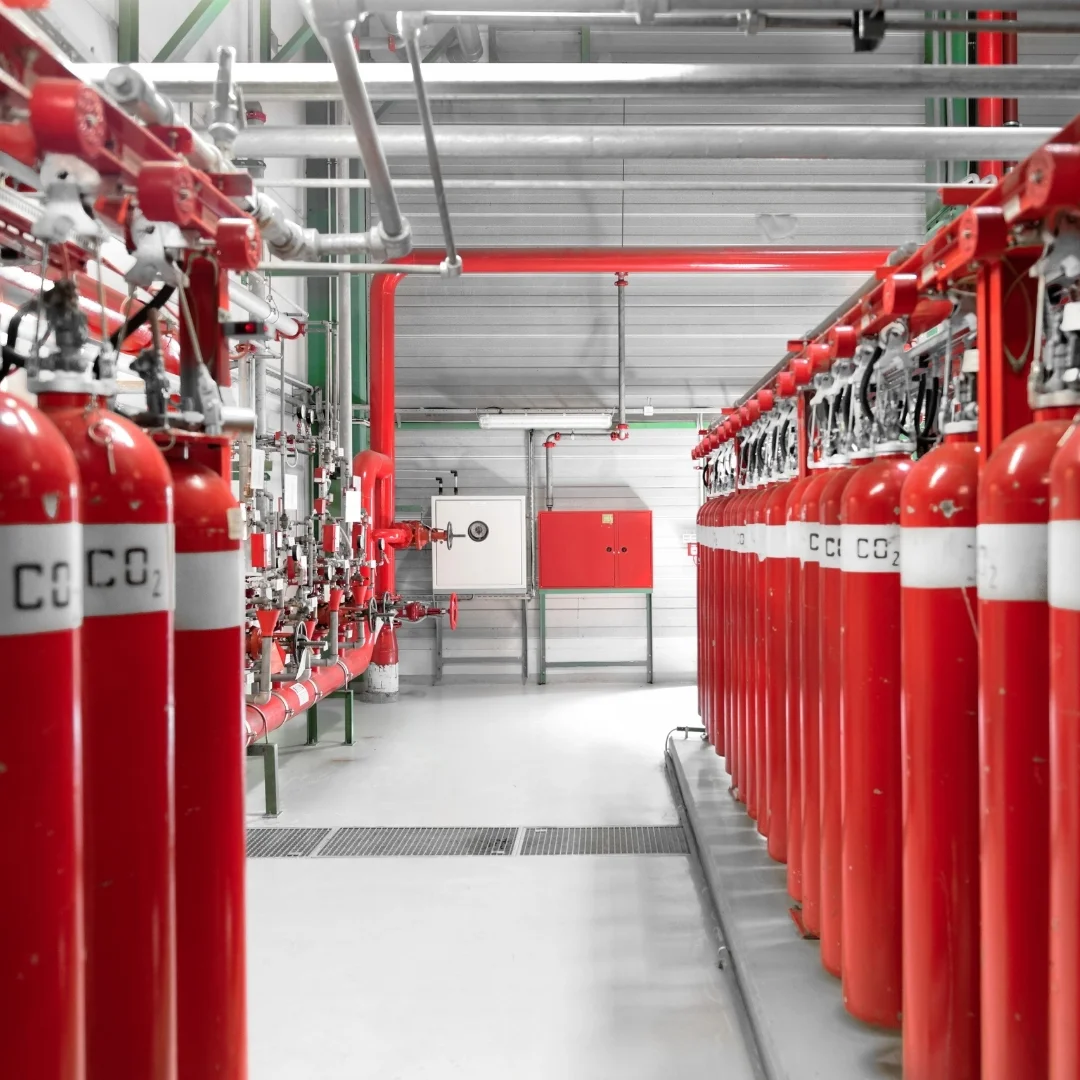 Rows of large red CO2 fire suppression cylinders connected to a manifold system and overhead piping in an industrial utility room.