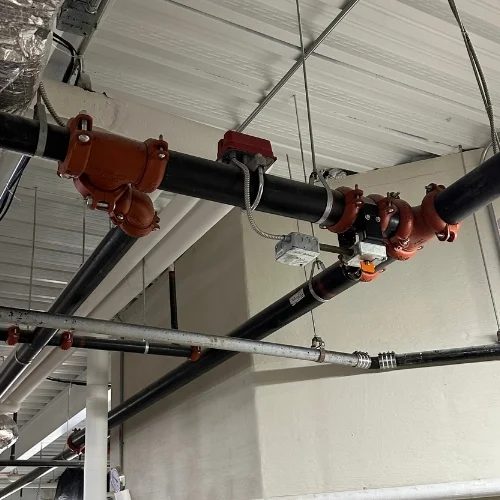 Black steel fire sprinkler main piping suspended from a ceiling, featuring orange grooved mechanical couplings and an electronic waterflow alarm switch with metal conduit.