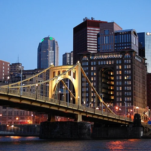 Evening view of the illuminated yellow Roberto Clemente Bridge (Sixth Street Bridge) spanning the Allegheny River, with the downtown Pittsburgh, Pennsylvania skyline rising in the background under a darkening sky.