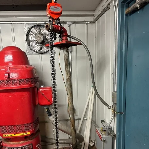 A large red vertical turbine fire pump motor standing next to a vertical shaft assembly with an overhead chain hoist hoist in a pump house utility room.