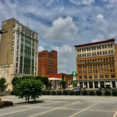 Ground-level view from an empty parking lot looking toward historic multi-story downtown commercial buildings, including a prominent white and green terracotta building on the left and a yellow brick building on the right, under a blue sky with puffy white clouds.